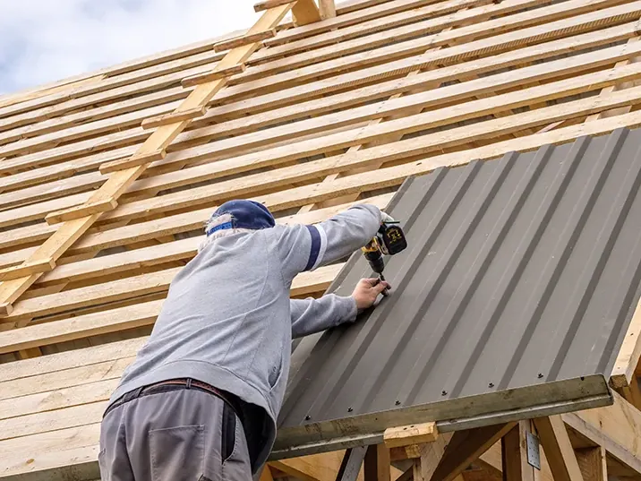 Worker fastening a gray metal roof panel to exposed wooden rafters
